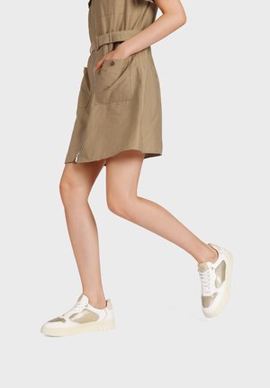 Gambe di una persona che indossa un vestito kaki con tasche frontali e sneakers bianchi, mostrata a metà passo su uno sfondo semplice.