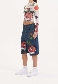 Blusa de mangas largas y transparente con gráficos florales; combinada con una falda de mezclilla hasta la rodilla con estampados de flores llamativos. Las zapatillas blancas completan el atuendo.