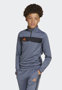 Top atlético gris con un panel en el pecho negro y acentos naranjas. Presenta un cuello alto y mangas largas, combinado con pantalones gris a juego.
