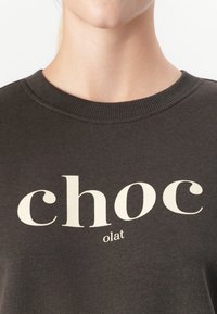 Primer plano de una persona que lleva una camiseta de cuello redondo marrón oscuro con la palabra "chocolat" impresa en letras grandes de color blanco sucio y partidas.