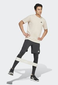 T-shirt beige chiaro a maniche corte con logo nero, abbinato a pantaloni sportivi neri e leggings, utilizzando una banda elastica bianca per l'esercizio.