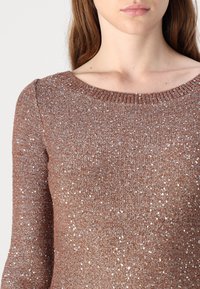 Femme portant un pull en tricot marron avec des sequins argentés, visible du cou jusqu'au milieu du torse sur un fond blanc uni.
