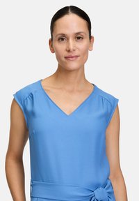T-Shirt mit V-Ausschnitt und kurzen Ärmeln aus hellblauem Stoff, der an den Schultern dezente Falten und eine gebundene Taillierung für Form und Passform aufweist.
