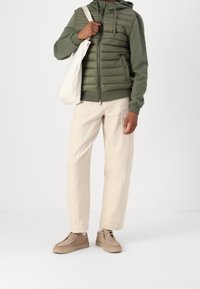 Pufjakke i olivengrøn med lynlås foran, kombineret med cremefarvede chinos og beige ruskindssko. Hvid tote-taske i hånden.