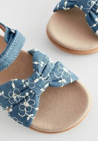 Siniset farkku sandaalit, joissa on valkoista kukkakirjontaa soljessa, pehmeä beige sisäpohja ja yksinkertainen hihnasuunnittelu.