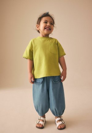 Usmívající se dítě stojí, má na sobě zelené tričko s krátkým rukávem a kapsou, volné modré kalhoty s volánovými kapsami a bílé sandály.