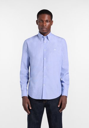 Uomo che indossa una camicia azzurro chiaro con piccolo logo bianco sul petto e jeans scuri, in piedi davanti a uno sfondo bianco uniforme.