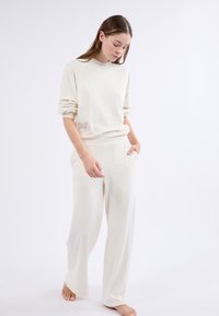 El conjunto de ropa de casa en color crema presenta una camiseta de ajuste relajado con manga larga y puños acanalados, junto con pantalones de pierna ancha a juego que cuentan con bolsillos laterales.