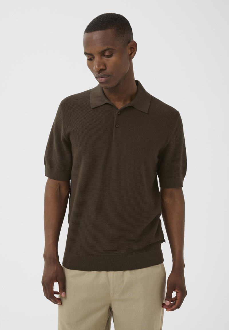 Homme portant un polo marron à manches courtes et un pantalon beige, debout les yeux baissés sur un fond clair uni.