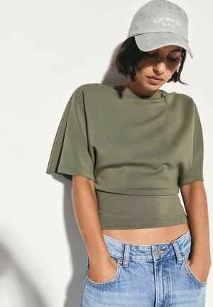 Femme portant un crop top vert olive, un jean bleu clair et une casquette grise avec le texte "Montmartre Paris", regardant vers le bas les mains dans les poches.