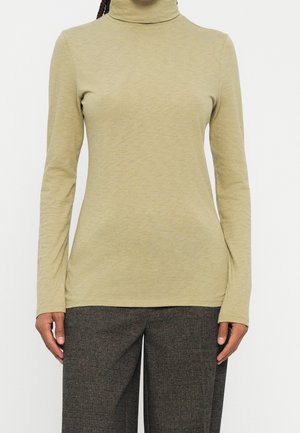 Beżowy golf z długim rękawem noszony z ciemnoszarymi, teksturowanymi spodniami, ręce luźno opuszczone wzdłuż ciała, na jednolitym białym tle.