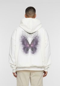 Sudadera blanca con un gran gráfico de mariposa morada en la parte posterior, con texto negro arriba y un ajuste holgado y cómodo. Tejido texturizado.
