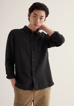 Młody mężczyzna o ciemnych włosach, ubrany w czarną koszulę zapinaną na guziki i beżowe spodnie, pozujący z jedną ręką na szyi na jednolitym tle.