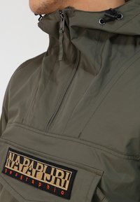 Uomo che indossa una giacca Napapijri verde oliva con mezza zip, colletto alto e tasca frontale con il logo del marchio.