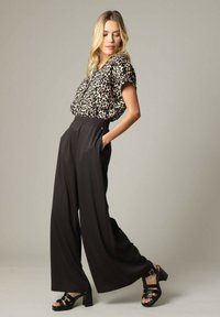 Blouse met luipaardprint en korte mouwen, gecombineerd met hoge zwarte wijde broek; model draagt zwarte hakken.