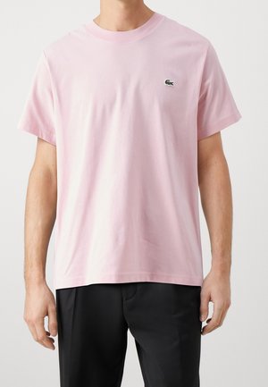 Mężczyzna noszący jasnoróżowy t-shirt z krótkim rękawem i małym zielonym logo krokodyla na lewej piersi, zestawiony z czarnymi spodniami.