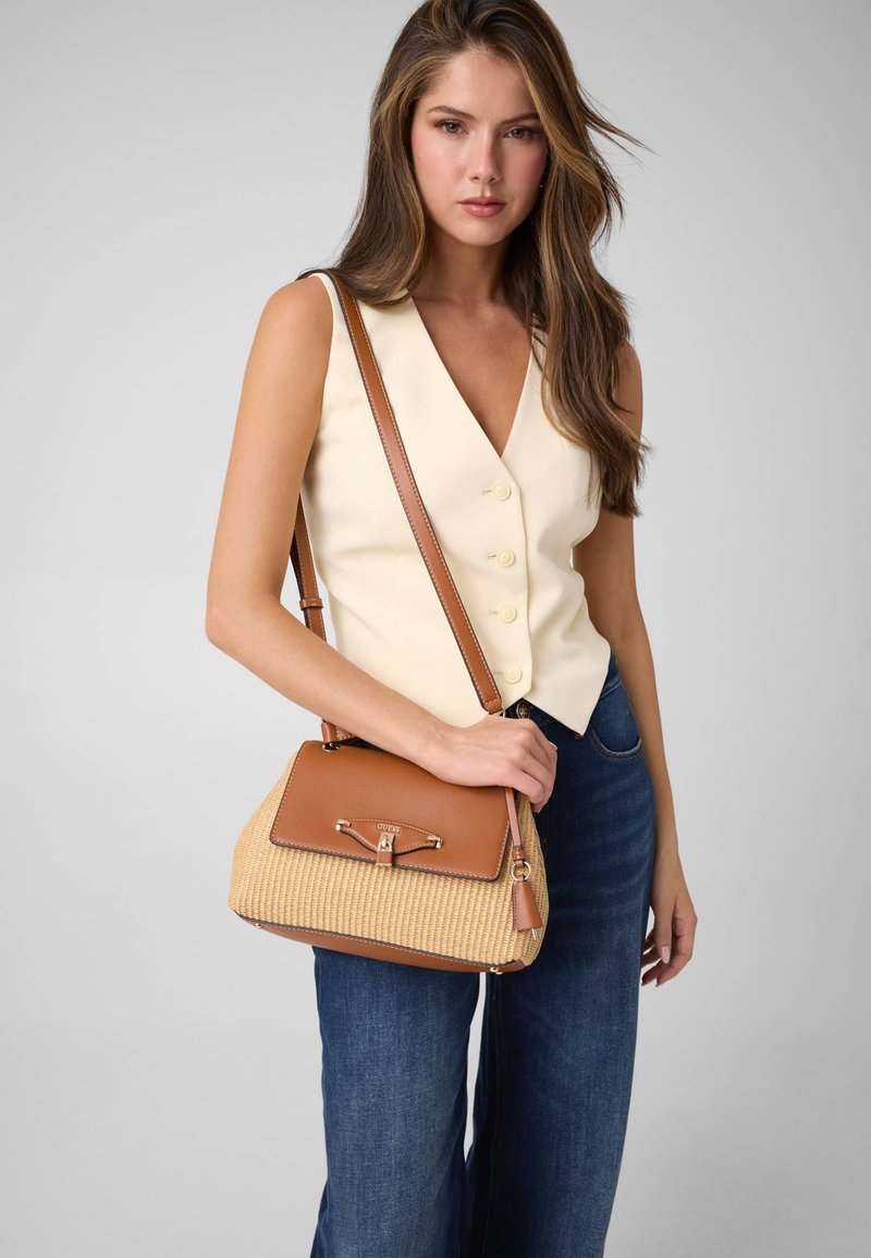 Femme portant un gilet sans manches crème et un jean bleu, tenant un sac à main marron et beige avec bandoulière sur fond uni.