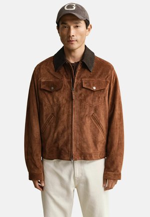 Homme portant une veste en daim marron avec un col foncé, un pantalon blanc et une casquette grise avec la lettre blanche "G" sur le devant, sur fond blanc.