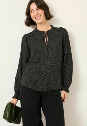 Schwarze Bluse mit Polka-Dots, langen Ärmeln, gerafftem Kragen und einem Bindeband vorne. Getragen mit schwarzen Hosen und einer grünen Handtasche.