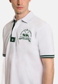 Camisa polo blanca con acentos verdes, con cuello y un logo bordado de dos palos cruzados y un caballo, tejido texturizado y mangas cortas.