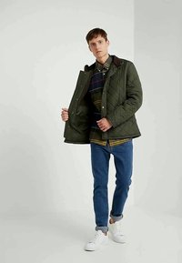 Ανδρας που φοράει πράσινη στιβαρή ζακέτα πάνω από καρό πουκάμισο, μπλε τζιν με μανίκια, και λευκά αθλητικά παπούτσια, στέκεται έναντι ενός απλού φωτεινού φόντου.