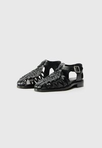 Sandalias de cuero negro con un diseño trenzado, lados abiertos y una correa de tobillo ajustable. Presentan un tacón plano y una punta redondeada.