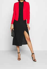 Kvinna iklädd en svart klänning med hög slits, röd blazer, svarta högklackade sandaler och håller en svart glittrande clutch.