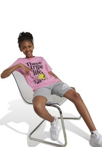 T-shirt rose à manches courtes avec imprimé graphique et texte noir, associé à un short adidas gris. Assis sur une chaise moderne et courbée. Baskets blanches.