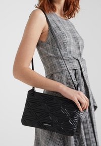 Žena v šedých kostkovaných šatech bez rukávů držící černou prošívanou kabelku Emporio Armani s geometrickými vzory.