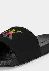 Sandale noire à enfiler avec semelle texturée et large bride en tissu arborant un logo Calvin Klein « CK » dégradé multicolore.