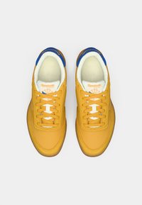 Par žutih Reebok Classic tenisica s bijelim mekanim unutarnjim dijelom i plavim naglascima, prikazanih odozgo na jedinstvenoj pozadini.