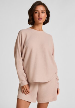 Femme portant un haut texturé à manches longues rose clair et un short assorti, posant devant un fond gris uni.