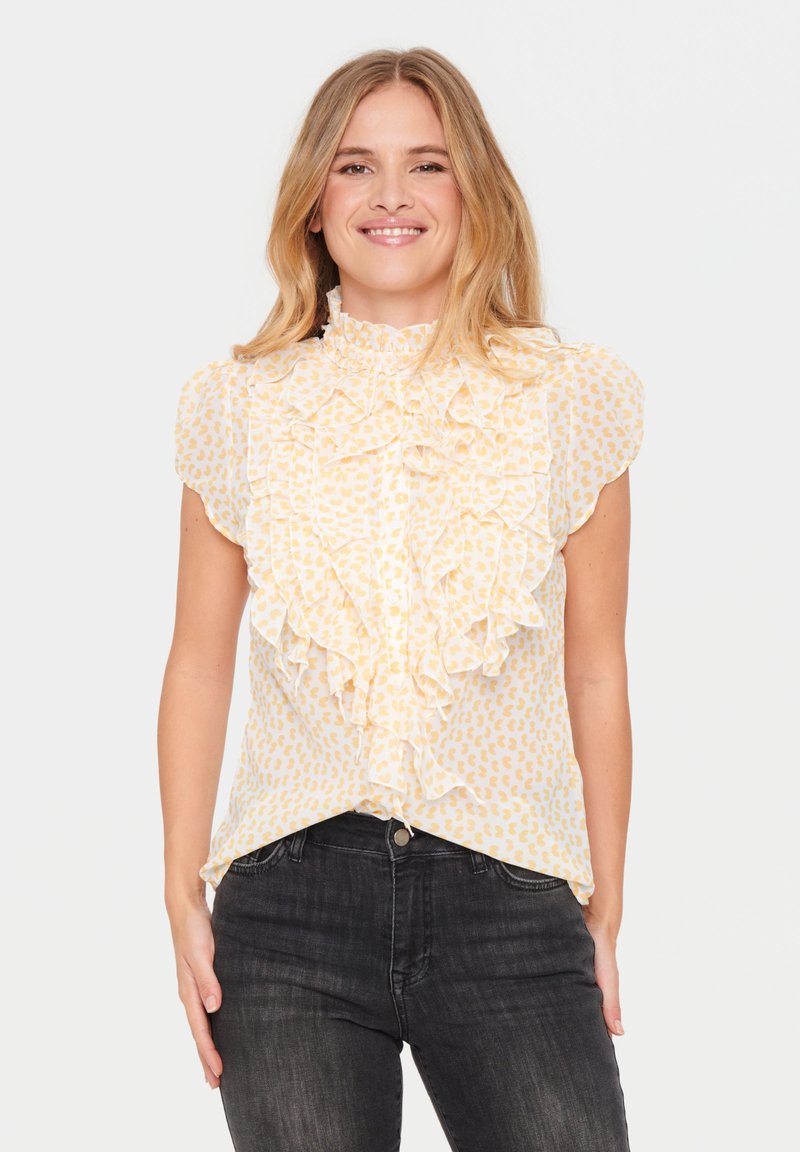 Lichtgele blouse met een gegolfd ontwerp, voorzien van een delicate print, korte mouwen en een hoge halslijn. Gepaarde met donkere jeans.