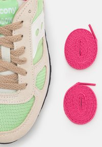 Sneaker Saucony Shadow in suede beige e rete verde, con lacci marroni; include due lacci tondi rosa su sfondo bianco.