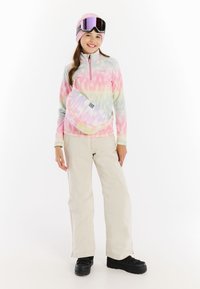 Fleeceová mikina s pastelovým tie-dye vzorem, béžové lyžařské kalhoty, růžová čepice a lyžařské brýle. Nosí bílou křížově umístěnou tašku s logem.