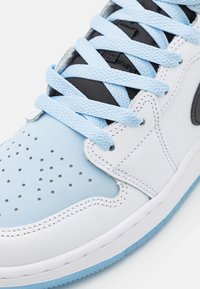 Baskets en cuir blanc avec des accents en daim bleu clair, pointe perforée, lacets bleus et sous-couche en maille noire. Semelle extérieure en caoutchouc transparent visible.