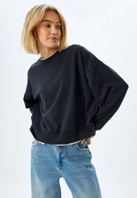 Navyblaues, oversized Sweatshirt mit langen Ärmeln, geripptem Halsausschnitt und cropped Saum. Weicher Stoff mit minimaler Textur, kombiniert mit hellblauen Jeans.