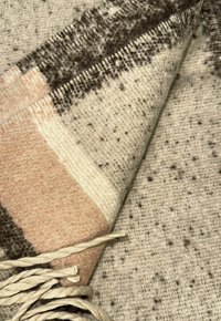 Couverture en mélange de laine avec une texture douce, des motifs géométriques beiges, bruns et roses modérés, avec des franges torsadées de couleur crème le long du bord.