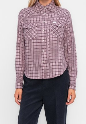 Kobieta w różowo-czarną, długim rękawem koszulę w kratę, z patkami na kieszeniach piersiowych oraz ciemne spodnie, na jednolitym tle.