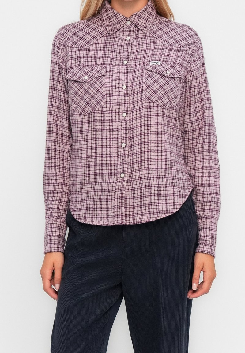 Femme portant une chemise à carreaux rose et noire à manches longues, boutonnée, avec poches poitrine à rabat et pantalon foncé sur un fond uni.