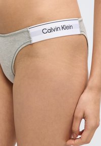 Γκρι βαμβακερό στρινγκ με λευκή ελαστική μέση που φέρει το λογότυπο "Calvin Klein" με μαύρα γράμματα. Ομαλή υφή και μινιμαλιστικός σχεδιασμός.