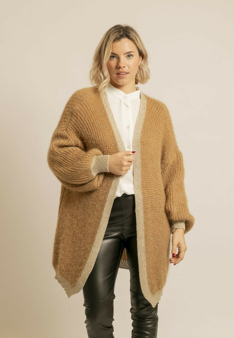 Cardigan en tricot brun avec une coupe loose, présentant une bordure argentée le long des bords. Le tissu semble doux et texturé, avec des épaules tombantes.