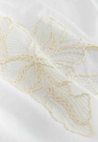 Gros plan sur un tissu blanc avec un motif floral brodé couleur crème et des détails de coutures texturées.