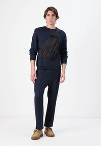 Zwarte schoudertas met een driehoekige vorm, over het lichaam gedragen over een marineblauwe sweatshirt en bijpassende broek. Bruine schoenen maken de look compleet.