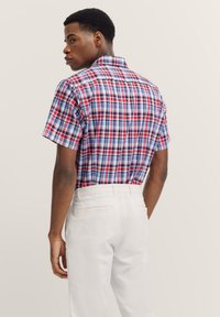 Kurzärmliges karierter Hemd in Rot, Blau und Weiß, mit Kragen und tailliertem Schnitt. getragen mit weißen Hosen, zeigt eine Rückansicht.