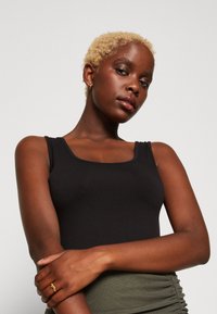 Black fitted tank top with a square neckline, smooth texture. Model's hands crossed, wearing a gold ring and a green ruched skirt.