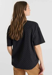 Camisa de manga corta negra con lunares blancos, ajuste relajado y dobladillo redondeado. Tejido texturizado con un acabado suave.