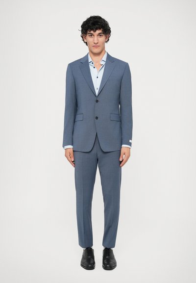 Traje ajustado azul claro con blazer de una sola botonadura y pantalones de frente plano. Camisa de vestir blanca con detalles en el cuello y los botones. Zapatos negros.
