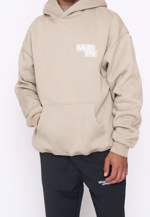 Personne portant un sweat à capuche beige avec le texte « Multiply » et un pantalon noir avec le texte blanc « Multiply Apparel » sur la cuisse, mains relâchées le long du corps.