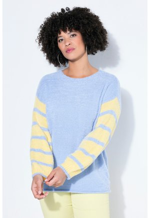 Kobieta o kręconych włosach, ubrana w jasnoniebieski sweter z żółto-niebieskimi paskami na rękawach oraz w jasnopomarańczowe spodnie, stojąca z dłońmi splecionymi.